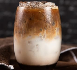 Ice coffee, toppings by floating foam milk and burned sugar in a tall of clear glass on dark gray real wood and chopping board. Decoration with coffee bean. Popular drink to quench thirst to cool down